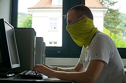 Das Bild zeigt einen Studenten am Computer mit improvisierter Maske.