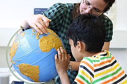Das Bild zeigt einen blinden Jungen, welcher einen taktilen Globus erkundet. Neben ihm steht eine erwachsene Frau. Kind neben einem Globus