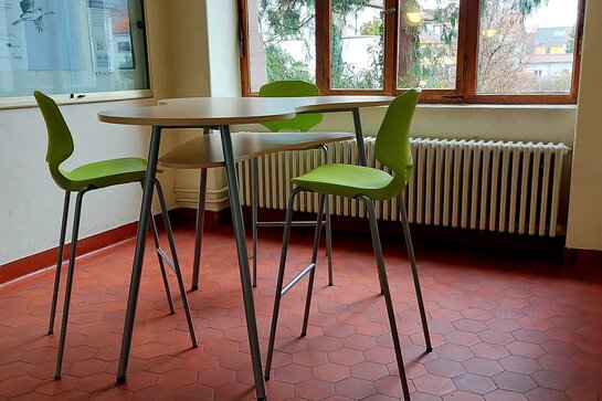 Abgebildet ist ein Steharbeitsplatz im Altbaufoyer. Grüne Stühle in Barhockerhöhe ermöglichen auch sitzendes Arbeiten. Durch das Fenster hat man einen Blick auf einen alten Baum