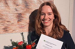 Porträt von Lea Hornscheidt. Sie steht vor ihrem Gewinnerbild. Sie hält Blumen und die Urkunde in der Hand.