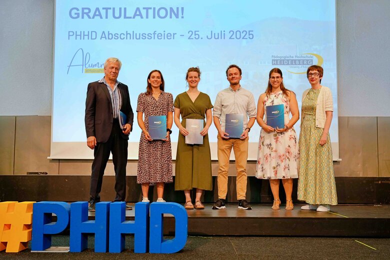 Zu sehen sind Alumni, die auf einer Bühne stehen und ihre Bestehensbriefe in PHHD-Mappen hochhalten. Gemeinsam mit Rektorin Vach & Prorektor Dammer (a.D.), welche ihnen die Mappen überreicht haben.