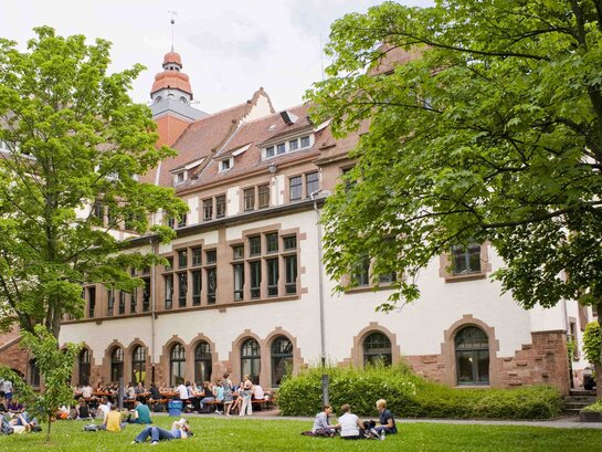 Das Bild zeigt den Innenhof und einen Flügel des Altbaus der PH Heidelberg. Im Innenhof auf dem Gras sitzen Studierende.Es ist Sommer.