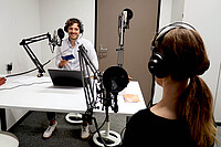 Auf dem Bild ist primär Max Wetterauer zu sehen. Er sitzt im Podcast-Studio der Hochschule und schaut lächelnd zu einer Frau, die man vorne rechts von hinten sieht. Beide tragen Kopfhörer. Copyright: PH Heidelberg