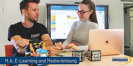 Zwei Studierende sitzen an einem Tisch. Vor Ihnen ein Laptop und Robitik. Unten Links ein Schriftzug: M.A. E-Learning und Medienbildung