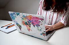 Auf dem Symbolbild ist primär ein Laptop von hinten zu sehen. Auf der Rückseite des Laptops klebt eine große, bunte Weltkarte. An dem Laptop sitzt eine Frau, deren Gesicht nicht zu sehen ist. Copyright: PH Heidelberg