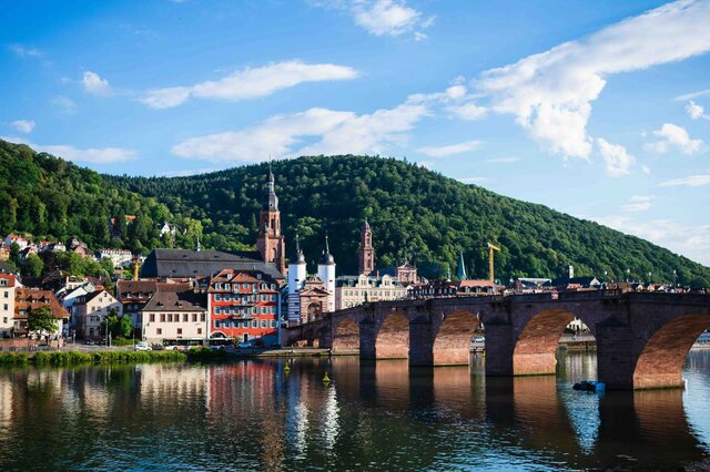 Zu sehen ist eine Ansicht der Heidelberger Altstadt, im Vordergrund die alte Brücke über den Neckar.