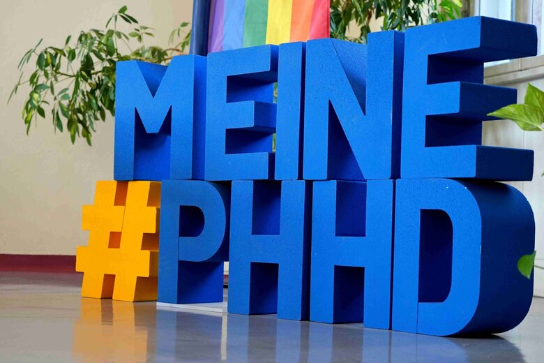 Der Hashtag #MeinePHHD in großen, einzelnen Buchstaben. Dahinter die Diversity-Flagge der Pädagogischen Hochschule Heidelberg.