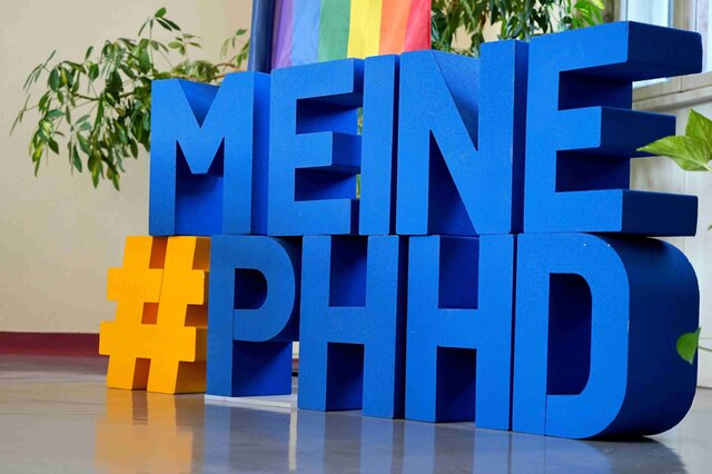 Der Hashtag #MeinePHHD in großen, einzelnen Buchstaben. Dahinter die Diversity-Flagge der Pädagogischen Hochschule Heidelberg.