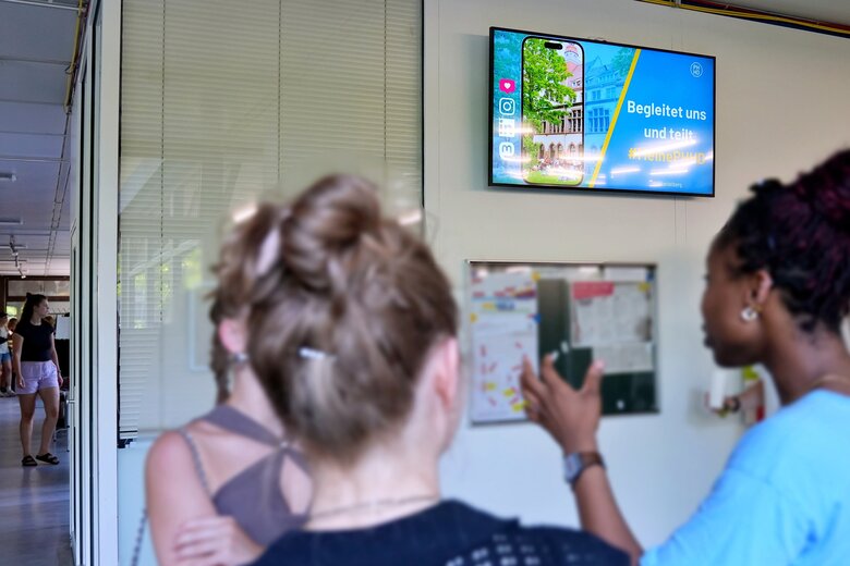 Eine Gruppe von Studierenden steht vor dem digitalen Schwarzen Brett im Neubau. Der Fokus liegt auf dem Bildschirm; die Studierenden sind unscharf. Das gilt auch für den Neubauflur, den man links im Bild sieht.