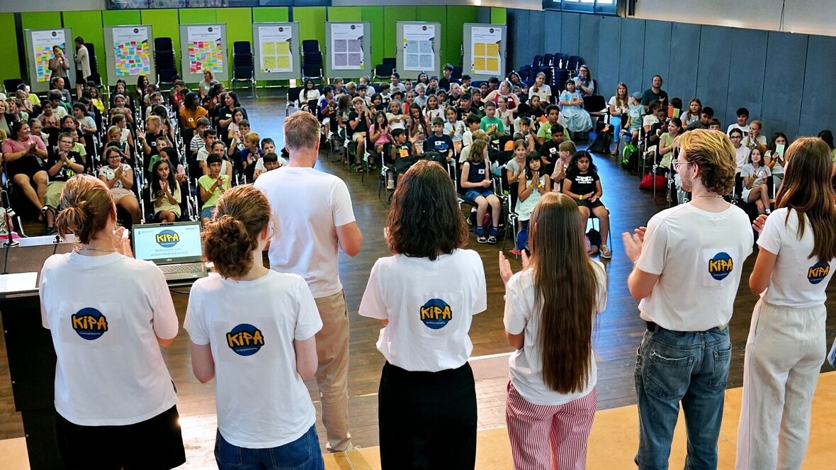Blick von der Bühne der Festhalle ins volle Plenum. Man sieht das studentische Team der Kinderparlamente von hinten und blickt auf zahlreiche Kinder, die aufmerksam zu hören.