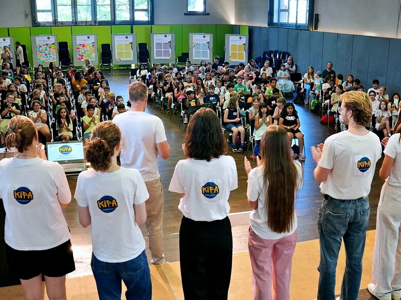 Blick von der Bühne der Festhalle ins volle Plenum. Man sieht das studentische Team der Kinderparlamente von hinten und blickt auf zahlreiche Kinder, die aufmerksam zu hören.