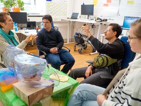 Das Bild zeigt einen Ausschnitt der Fortbildung. Vorne sind Modelle der weiblichen und männlichen Geschlechtsorgane auf einem Tisch, dahinter die Qualifizierungsteilnehmenden mit der Kursleiterin Kaya Schilling.