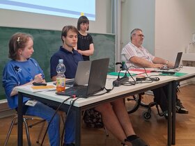 Das Bild zeigt Louisa Kabbe und Susann Bensch bei ihrer Modulprüfung. Sie sitzen bzw. stehen vor einer Tafel in einem Seminarraum der PH Heidelberg. Mit dabei auf dem Bild sind FSJ-Kraft Carl Hoffmann sowie Bildungsfachkraft Helmuth Pflantzer.