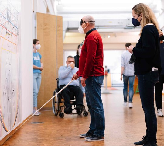 Zwei Personen betrachten ein großes Bild in einem Museum. Eine davon trägt eine  Augenbinde und hält einen Blindenstock.