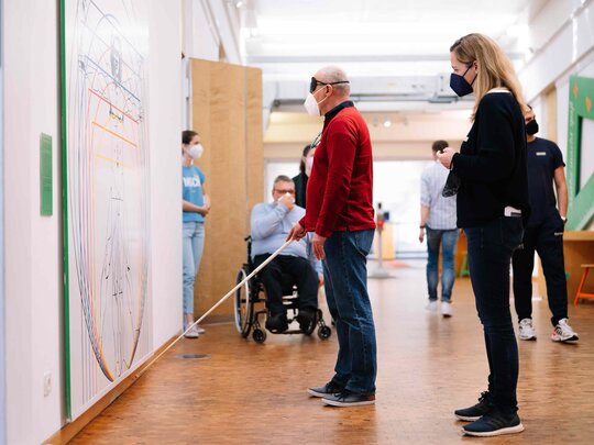 Zwei Personen betrachten ein großes Bild in einem Museum. Eine davon trägt eine  Augenbinde und hält einen Blindenstock.