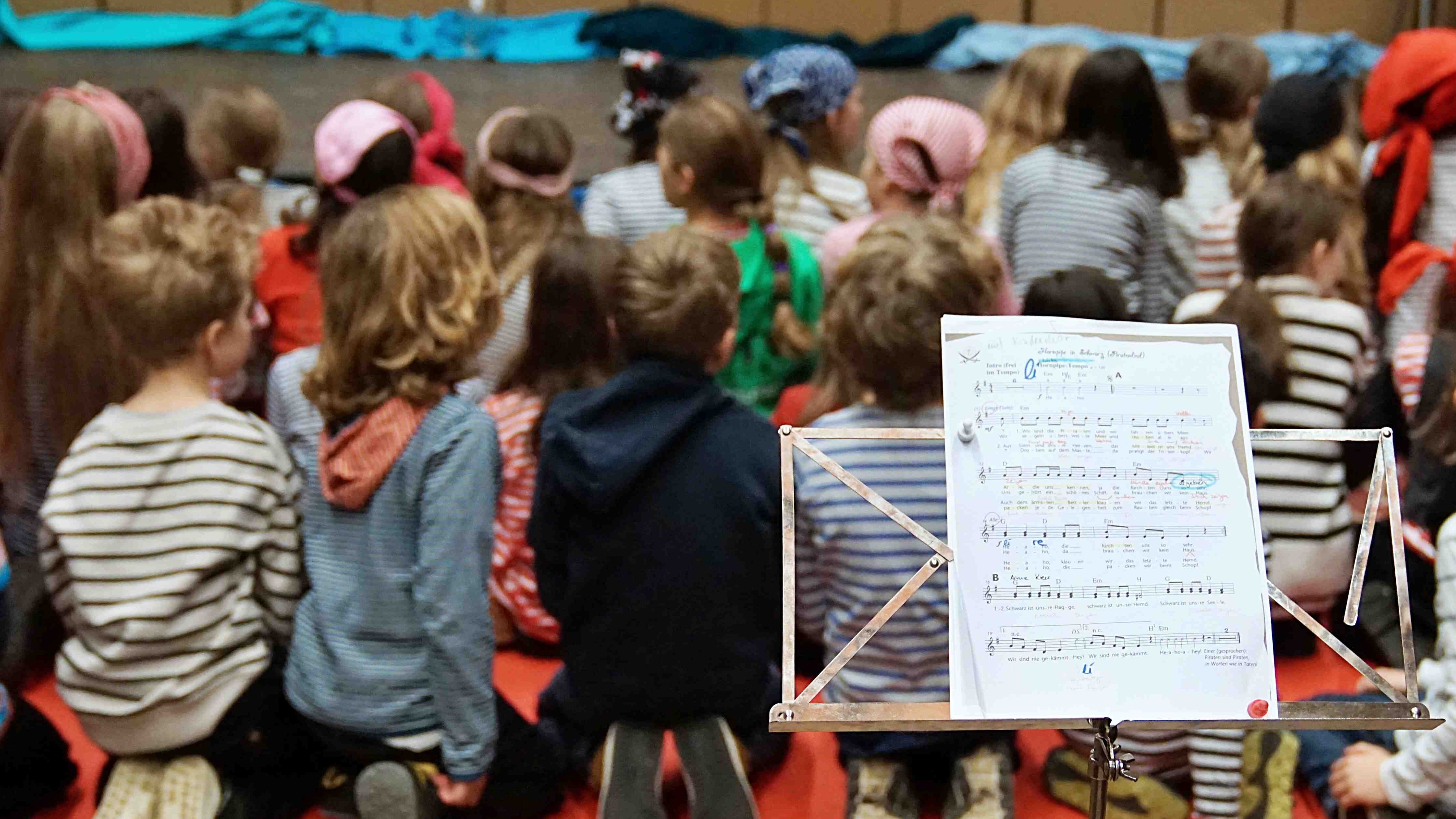 Eine Gruppe Grundschulkinder sitzt vor einer Bühne, im Vordergrund steht ein Notenständer mit Noten.
