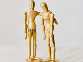 Zwei Holzfiguren (Zeichenpuppen) sind abgebildet. Eine hat eine traurige Körperhaltung, die andere scheint sie zu trösten und legt den Arm um die Schulter. Symbolbild für Erste Hilfe bei mentalen Krisen