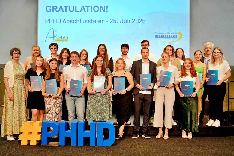 Zu sehen sind Alumni, die auf einer Bühne stehen und ihre Bestehensbriefe in PHHD-Mappen hochhalten. Gemeinsam mit Rektorin Vach & Prorektor Dammer (a.D.), welche ihnen die Mappen überreicht haben.