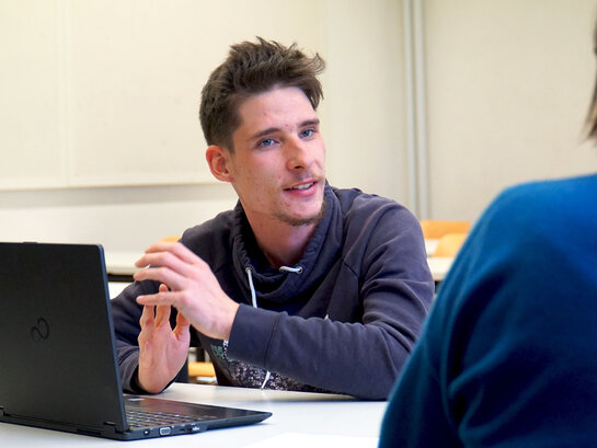 Das Bild zeigt eine Person am Notebook. Sie spricht mit einer Person, die nur von hinten zu sehen ist.