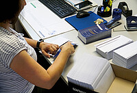 Das Bild zeigt eine Frau, die an einem Schreibtisch sitzt und die Einladung der Eröffnung des AW-ZIB in Umschläge steckt. Copyright: Pädagogische Hochschule Heidelberg