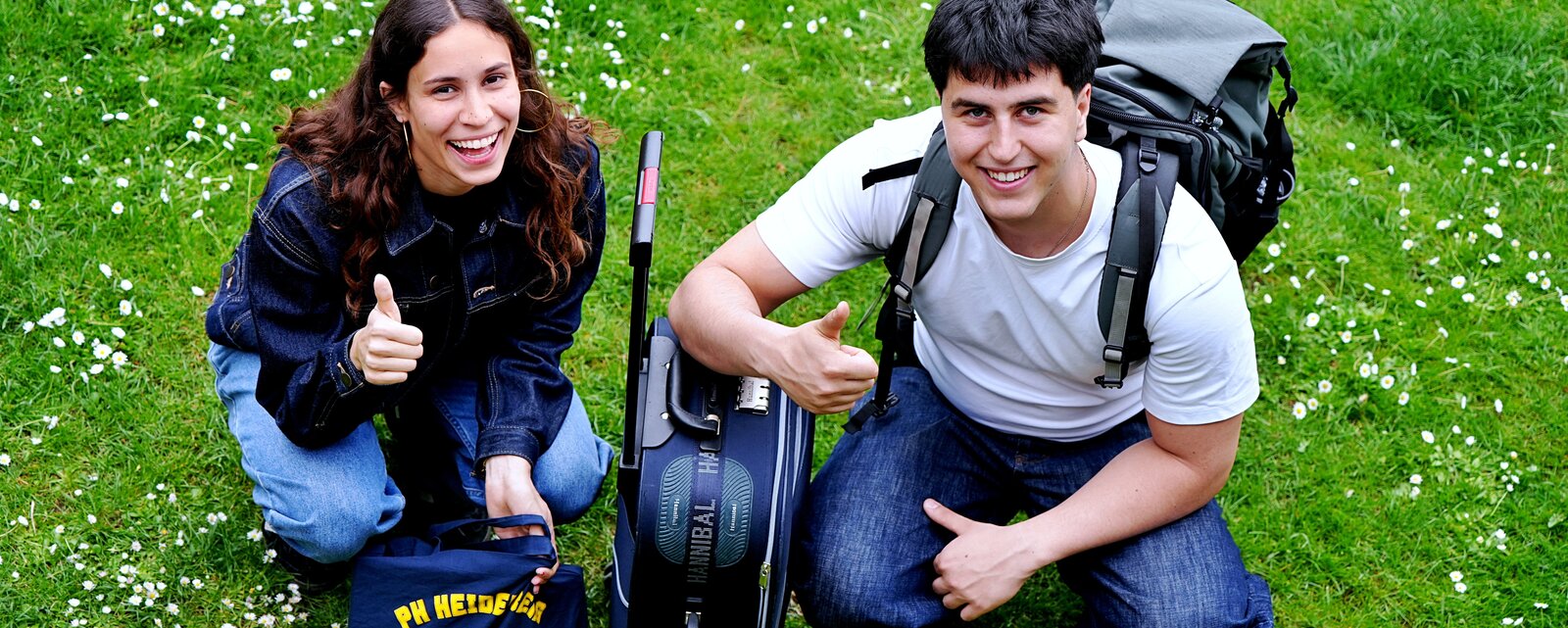 Zwei Menschen sitzen im Gras mit Rücksäcken und lachen in die Kamera