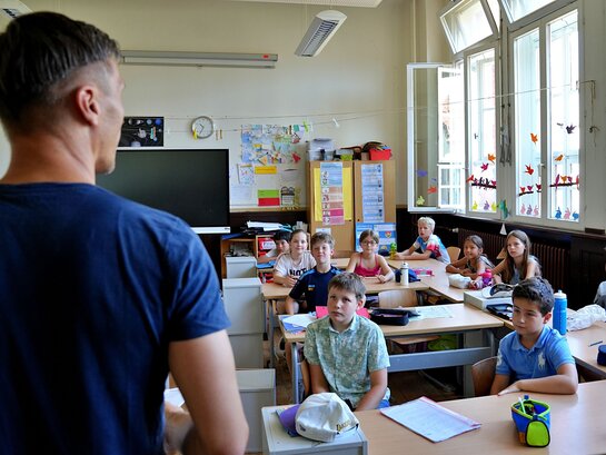 Das Foto zeigt eine stehene Person von hinten. Im Hintergrund sitzen viele Menschen und schauen auf die stehende Person.