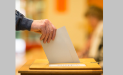 Das Foto zeigt eine Hand die einen Zettel in eine Wahlurne wirft.