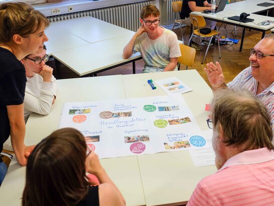 Das Bild zeigt eine Gruppe von Menschen, die miteinander sprechen. Sie sitzen oder stehen um einen Tisch herum. Auf dem Tisch liegt ein FlipChart-Papier, das bunt gestaltet ist. Darauf ist unter anderem zu lesen: Handlungsfelder im Quartier.