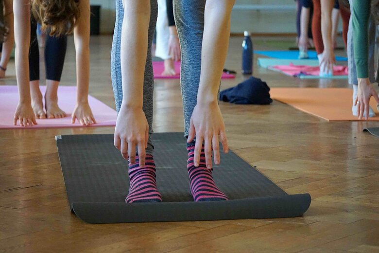 Zu sehen sind Fingerspitzen, die zu einer Matte auf dem Boden zeigen, es sieht aus als machen die Menschen Yoga oder Ähnliches. Dies ist ein Beispiel für den Hochschulsport.