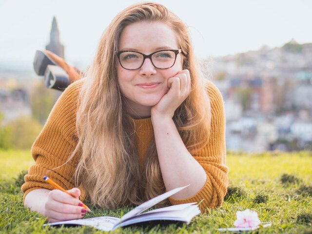 Eine versonnen lächelnde junge Frau liegt auf einer Wiese, vor sich ein Notizheft, in der Hand einen Stift.