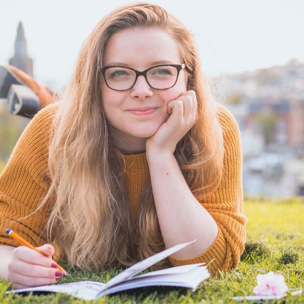 Eine versonnen lächelnde junge Frau liegt auf einer Wiese, vor sich ein Notizheft, in der Hand einen Stift.