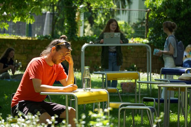 Zu sehen sind Studierende im Keplerhof des Altbaus der Pädagogischen Hochschule Heidelberg, welcher von PHeelGood eingerichtet wurde. Ein Student sitzt im Vordergrund, zwei Studierende stehen im Hintergrund an einem Stehtisch. 