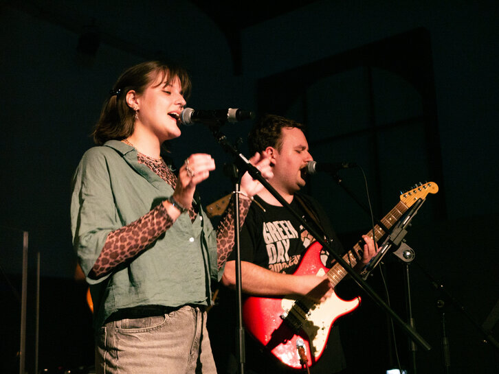 Eine junge Sängerin singt mit viel Energie ins Mikro, im Hintergrund steht ein Gitarrist, welcher die Zweitstimmen dazu beisteuert.