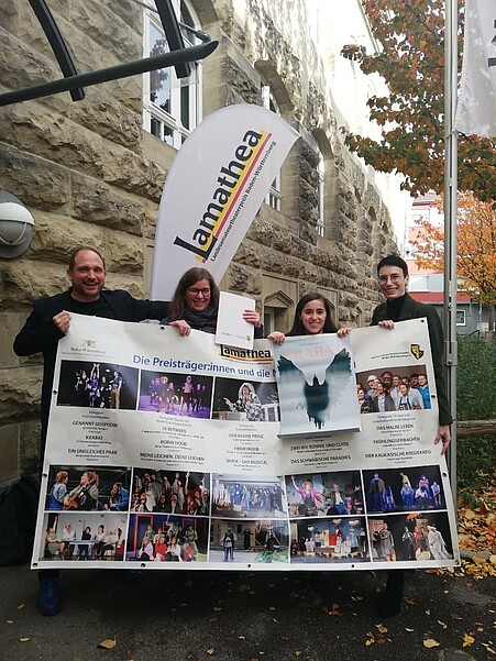 Teilnehmende, technische Leitung und Regie von Krabat stehen mit einem Banner der Lamathea Gewinner vor einem Steinhaus