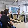 Das Bild zeigt Studierende in der GIS-Station der PH Heidelberg: Eine junge Frau steht vor dem Whiteboard und zeichnet auf eine Karte, die dort zu sehen ist; ein junger Mann zeigt von seinem Platz aus auf die Karte. Copyright: Pädagogische Hochschule Heidelberg.
