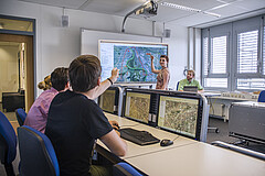 Das Bild zeigt Studierende in der GIS-Station der PH Heidelberg: Eine junge Frau steht vor dem Whiteboard und zeichnet auf eine Karte, die dort zu sehen ist; ein junger Mann zeigt von seinem Platz aus auf die Karte. Copyright: Pädagogische Hochschule Heidelberg.