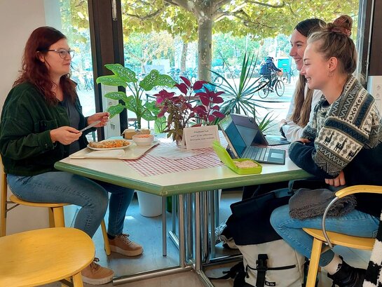 Studierende und Dozierende sitzen in der Neubaucafeteria am PHeelGood-Tisch und essen und reden gemeinsam.
