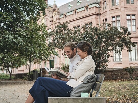 Eine junge Frau und ein junger Mann sitzen auf einer Bank im Park. Im Hintergrund sieht man unscharf die Bibliothek der Universität.