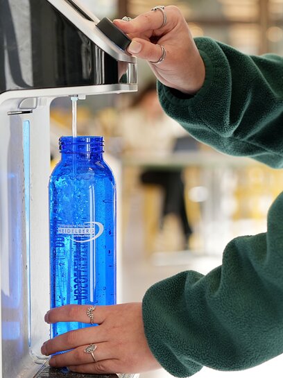 Eine blaue Wasserflasche der PHHD wird in einem Wasserspender mit Sprudelwasser gefüllt