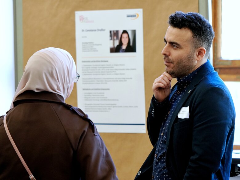Ein Frau und ein Mann stehen vor einem Plakat der DGFF-Tagung. Sie trägt Kopftuch und ist von hinten zu sehen. Er ist i Profil zu sehen und spricht gerade.