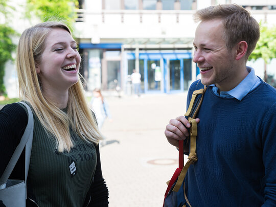 Eine Studentin und ein Student fröhlich lachend vor einem Hochschulgebäude
