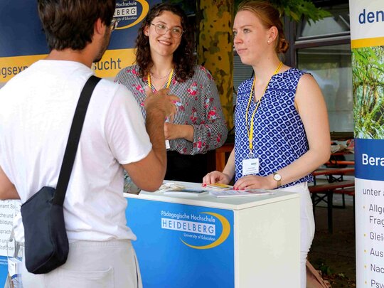Zwei studentische Tutorinnen stehen hinter einem Counter der PHHD. Davor steht ein junger Mann, der lebhaft mit den Händen spricht.