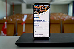 Das Symbolbild zeigt ein Handy, auf dem das Titelbild des QM-Berichts zu sehen ist. Das Handy steht auf einem zugeklappten Laptop in einem Hörsaal der Hochschule. Copyright: Pädagogische Hochschule Heidelberg.