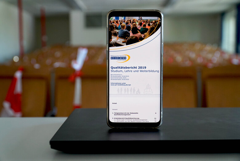 Das Symbolbild zeigt ein Handy, auf dem das Titelbild des QM-Berichts zu sehen ist. Das Handy steht auf einem zugeklappten Laptop in einem Hörsaal der Hochschule. Copyright: Pädagogische Hochschule Heidelberg.