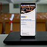 Das Symbolbild zeigt ein Handy, auf dem das Titelbild des QM-Berichts zu sehen ist. Das Handy steht auf einem zugeklappten Laptop in einem Hörsaal der Hochschule. Copyright: Pädagogische Hochschule Heidelberg.