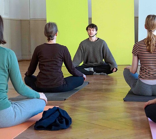 Mehrere Studierende sitzen auf Matten und praktizieren Yoga.