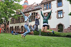 Das Foto zeigt drei springende Studierende vor dem Altbau der Hochschule.