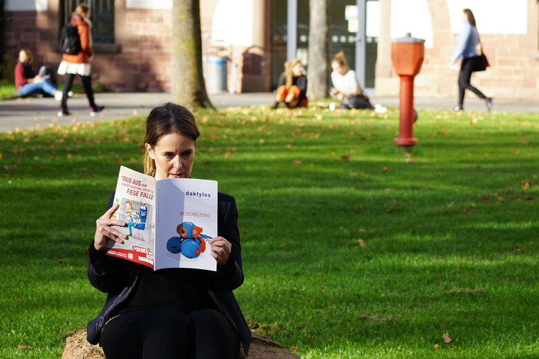 Frau sitzt im Innenhof der Hochschule und liest die aktuelle daktylos-Ausgabe. Im Hintergrund laufen Personen bzw. sitzen auf dem Gras.