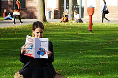 Frau sitzt im Innenhof der Hochschule und liest die aktuelle daktylos-Ausgabe. Im Hintergrund laufen Personen bzw. sitzen auf dem Gras.