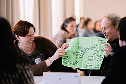 Zwei Frauen halten ein grünes Plakat in der Hand und blicken darauf. Im Hintergrund sieht man verschwommen andere KuLO-Teilnehmer:innen.